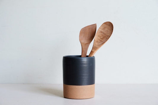 A cylinder utensil holder dipped half in black glaze, made of speckled stoneware.