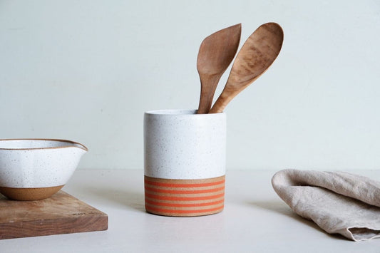 A cylinder utensil holder dipped half in white glaze, with rings of red glaze stripes at the bottom, made of speckled stoneware.